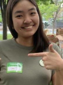 Phoebe is smiling and pointing to a nametag that says 'I share'. She has brown hair and a green shirt. 